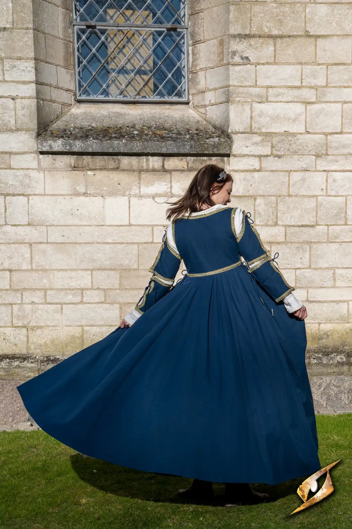 Dress Lucrezia, a stunning blue gown with golden trim, flowing gracefully in the breeze against a historic stone backdrop.