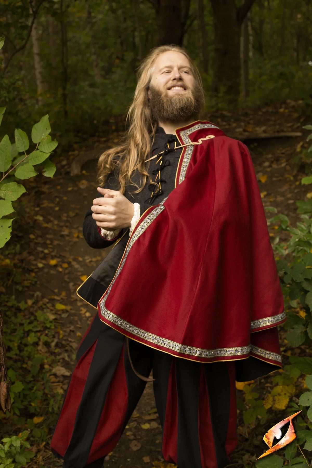 A man wearing Cape Aramis, a stylish red caplet with a high collar and gold trim, posing confidently in nature.