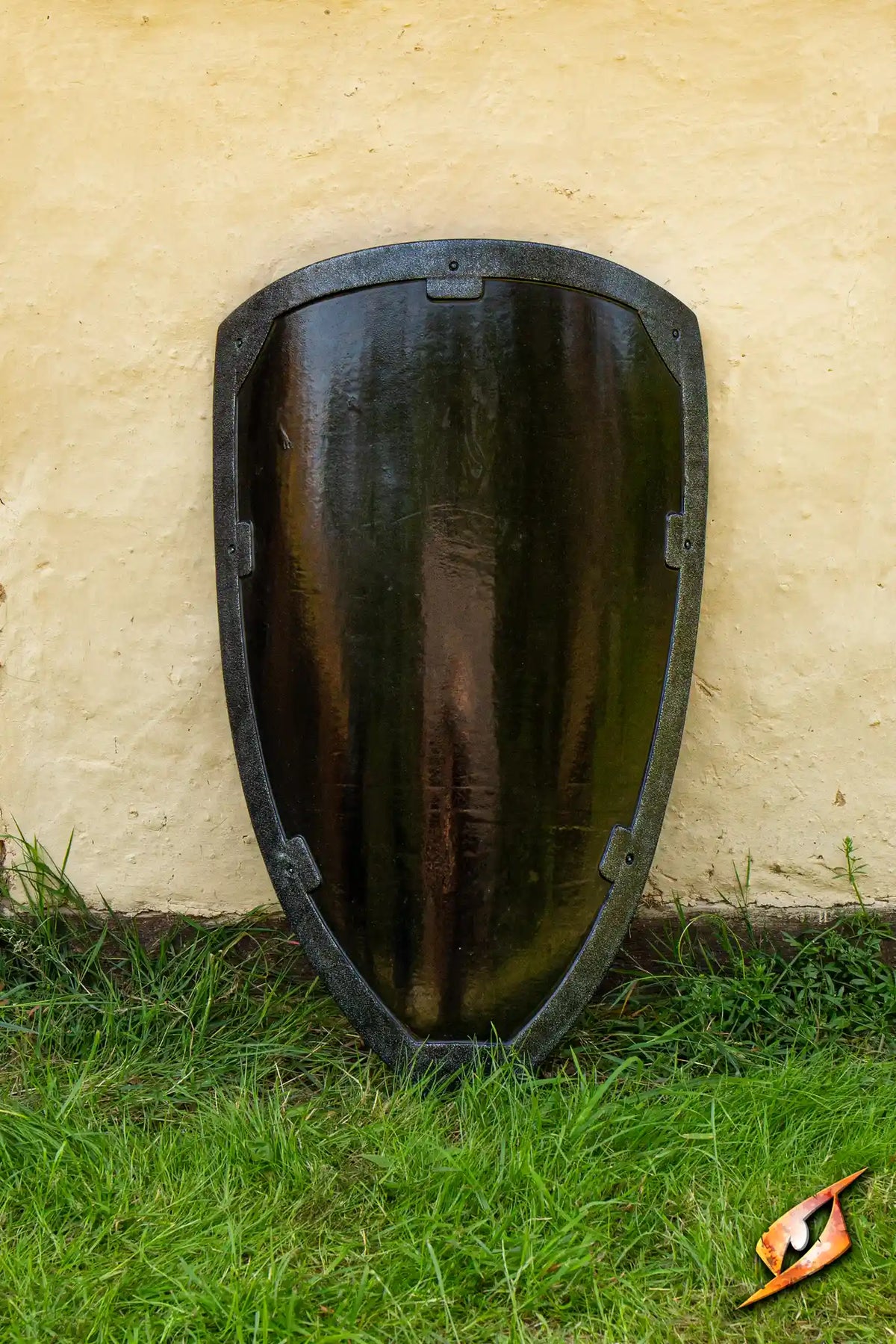 A Black Knight Shield displayed upright on grass against a textured wall, featuring a glossy black finish and tarnished metal rim.