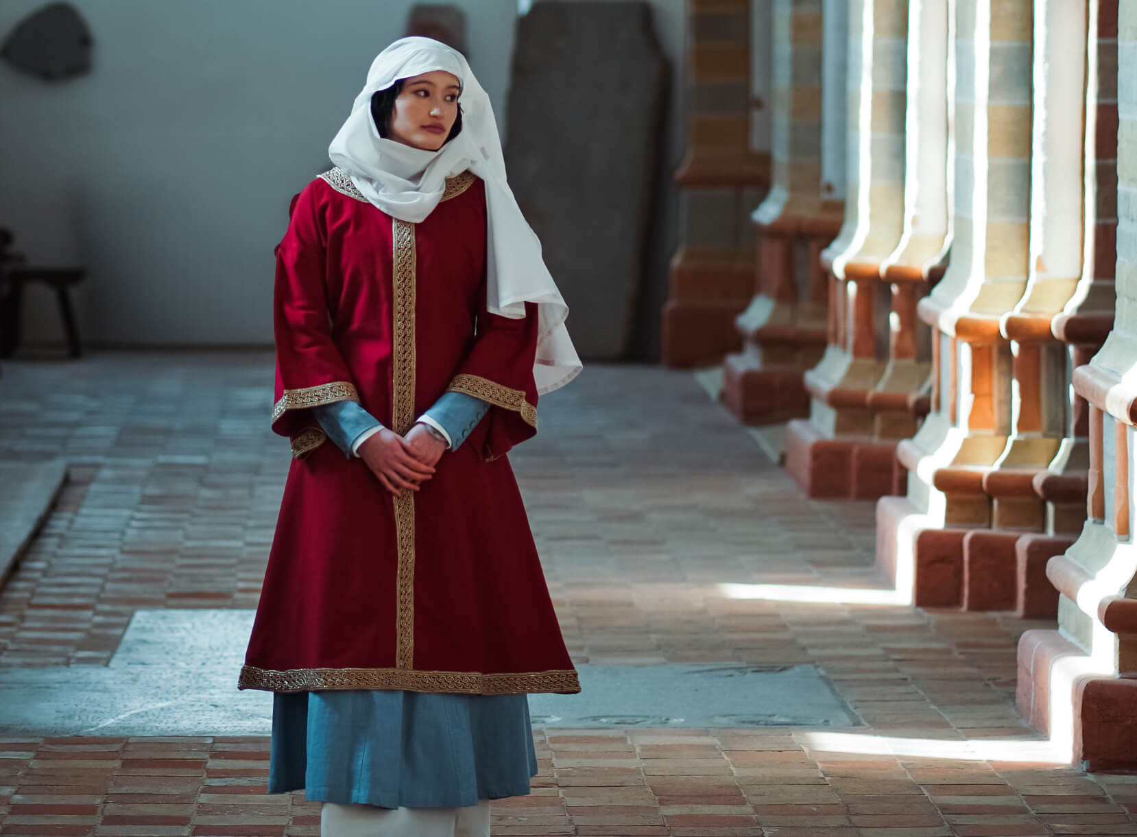 Woman in medieval red robe and white head wrap in a stone hall.