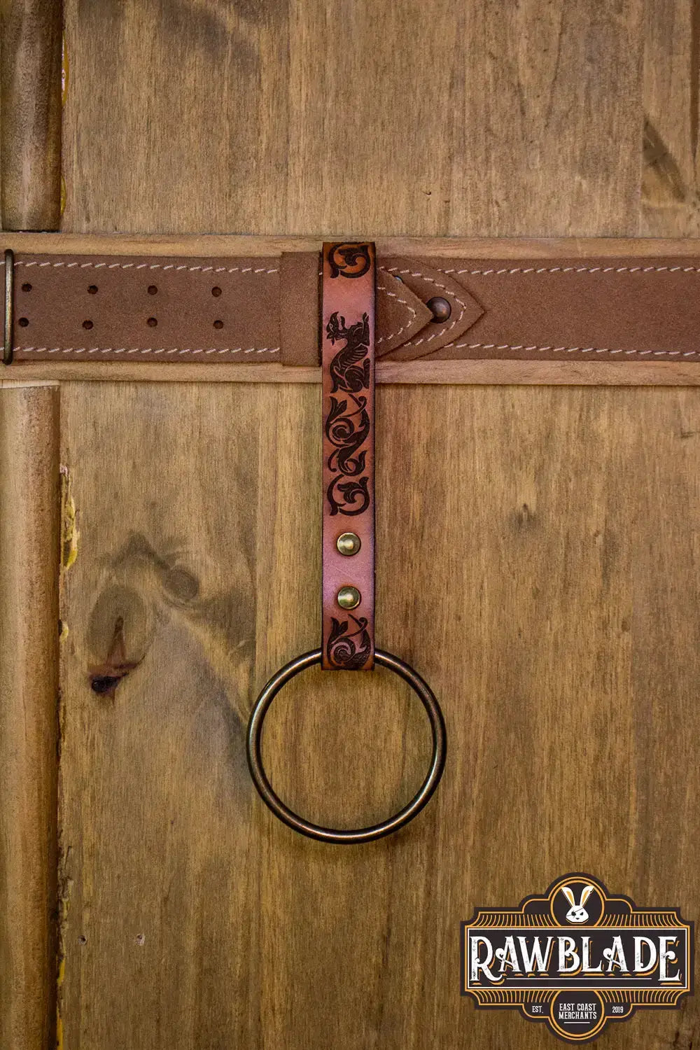 Aquitania Ring Holder with ornate laser engraving and gold rivets on a leather strap against a wooden background.