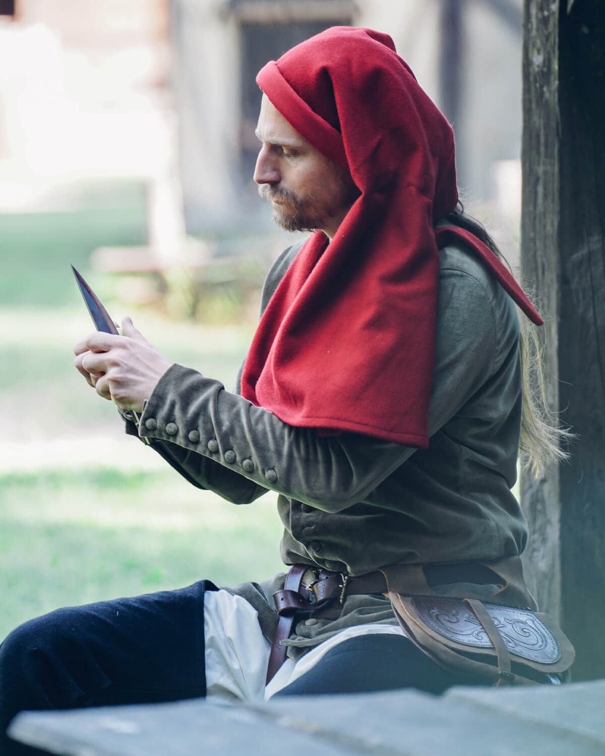 Man in medieval costume with red hood and brown belt, looking at dagger.