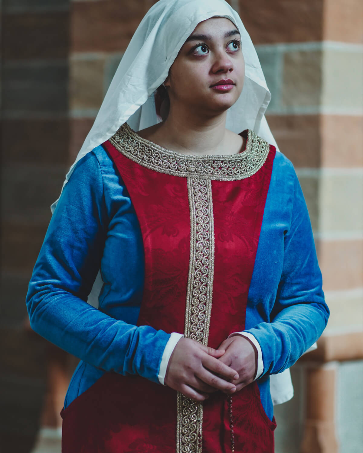 Woman in medieval blue and red dress with white head covering, hands clasped.