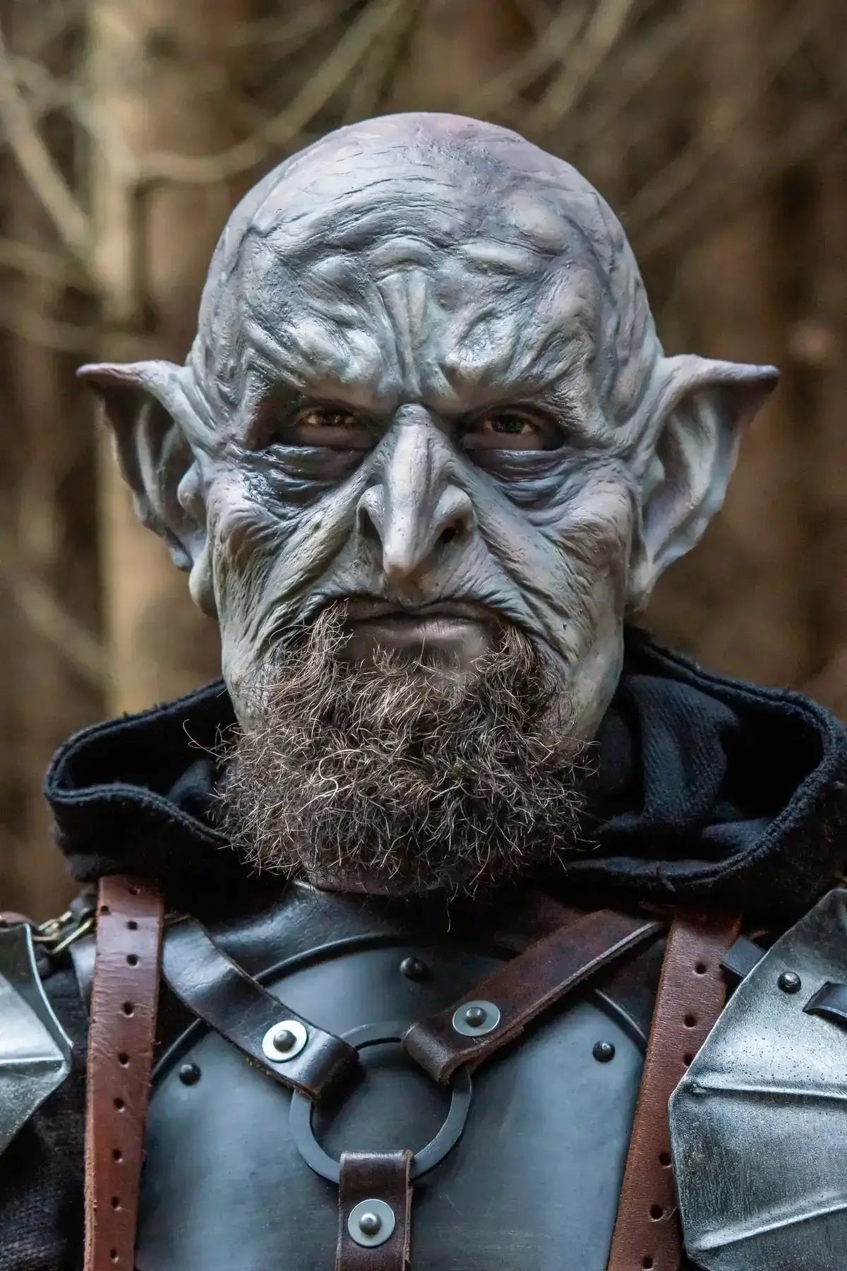 A close-up portrait of a person wearing a highly detailed fantasy costume, featuring a textured gray and silver orc-like mask with pointed ears, a brown beard, and dark armor with leather straps.