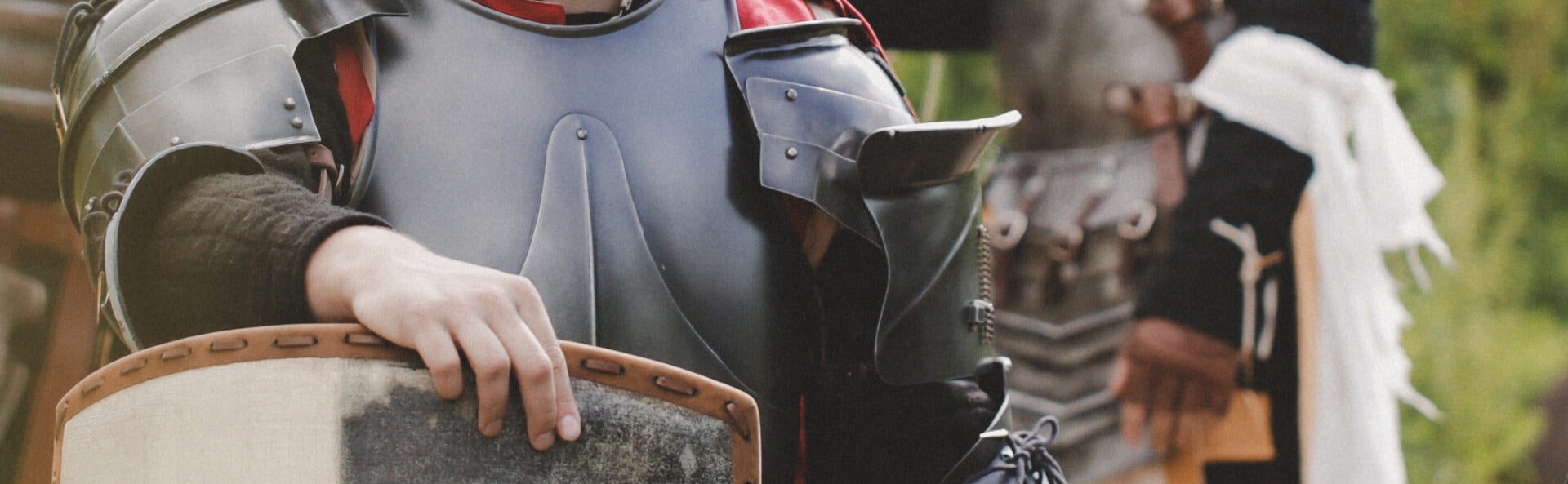 Person in medieval armor holding a shield with another person in similar attire in the background.