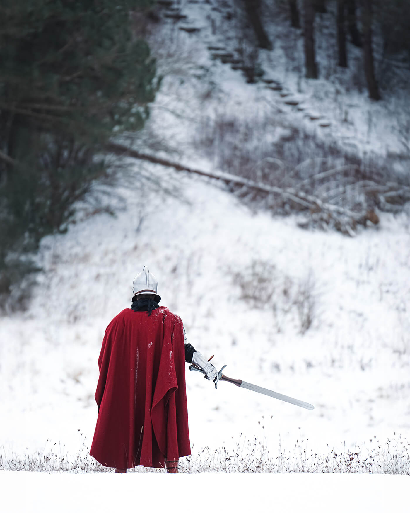 Person in a red cloak holding a sword walking through a snowy landscape