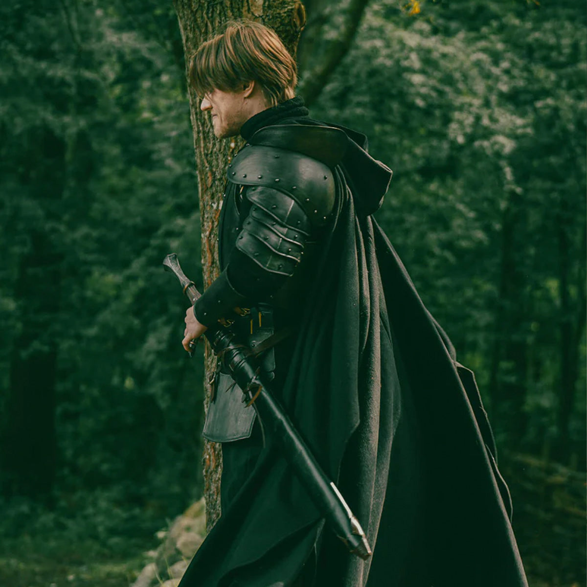 Man in a cloak standing by a tree with a sword, representing cloaks, capes & mantles: What’s the difference and how are they used today?