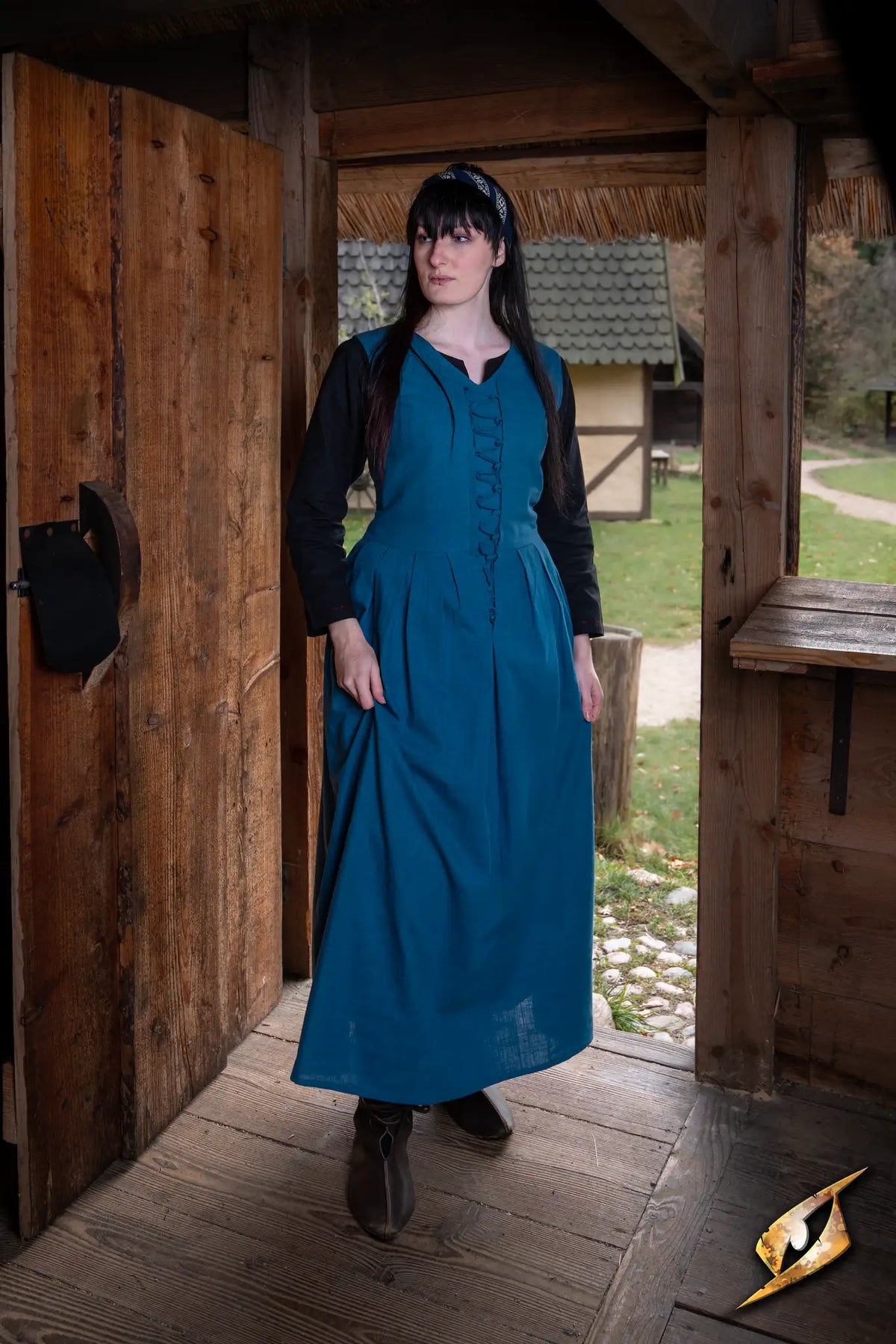 Model wearing the Adelina Dress, a medieval-style teal cotton dress, at a rustic outdoor setting.