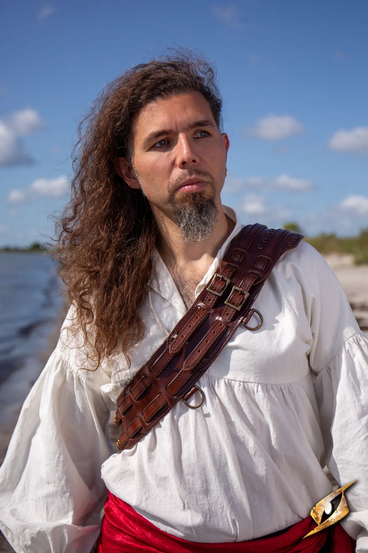 Man wearing a bandolier of many adventures, standing near the shore with a contemplative expression.