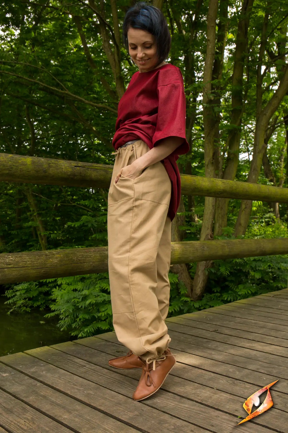 Woman wearing Basic Pants and a red top while standing on a wooden bridge in a lush green environment.