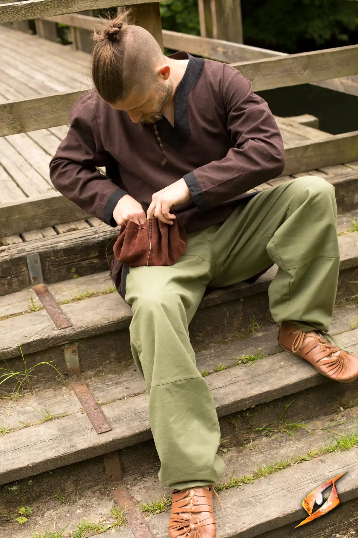 Man wearing Basic Pants sitting on wooden steps, adjusting a bag, dressed in a brown shirt and sandals.