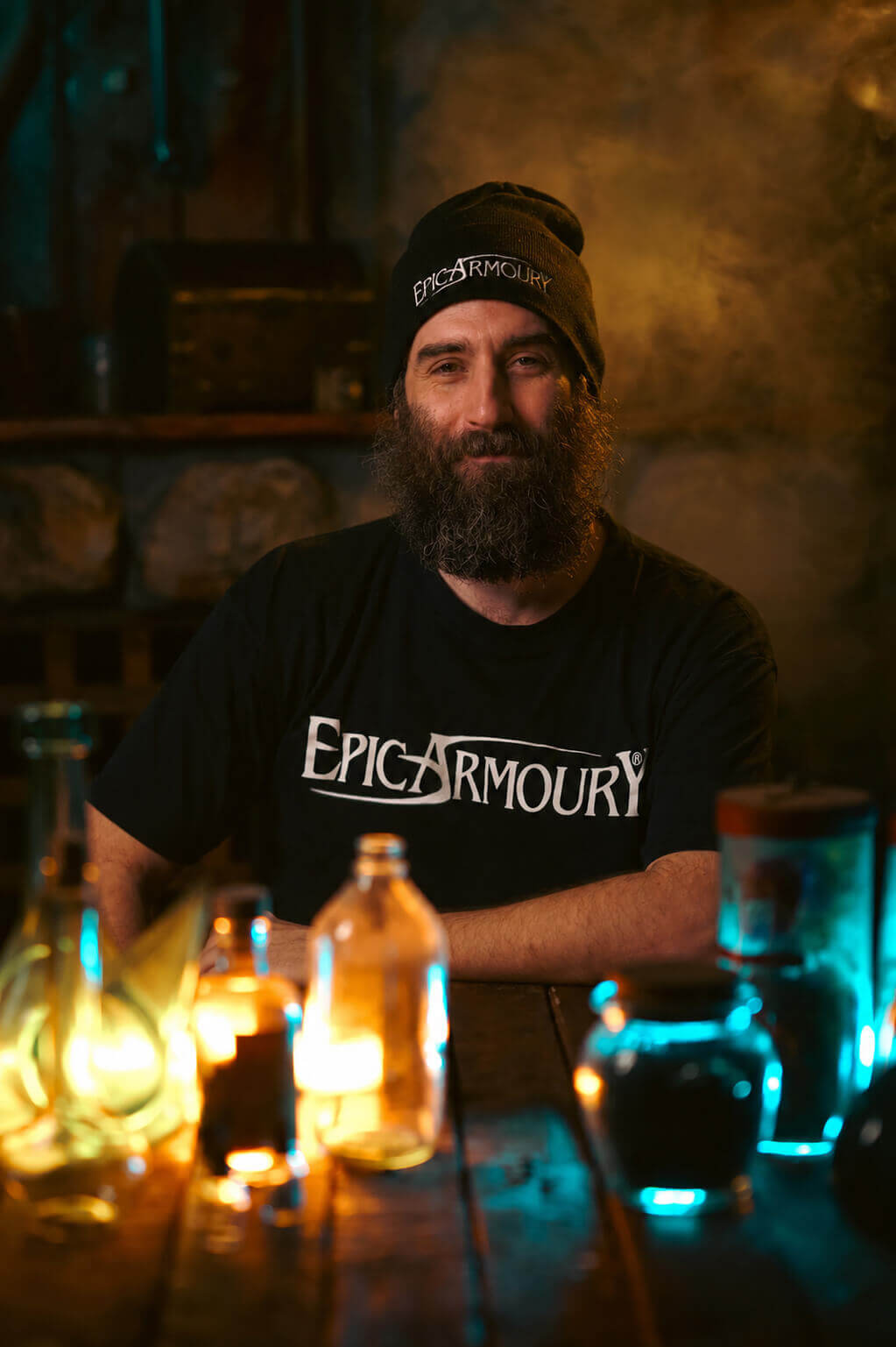 A bearded man wearing a black beanie and a black t-shirt with 'EPIC ARMOURY' printed in white across the chest, sitting at a wooden table with glowing bottles in a dimly lit, atmospheric setting.
