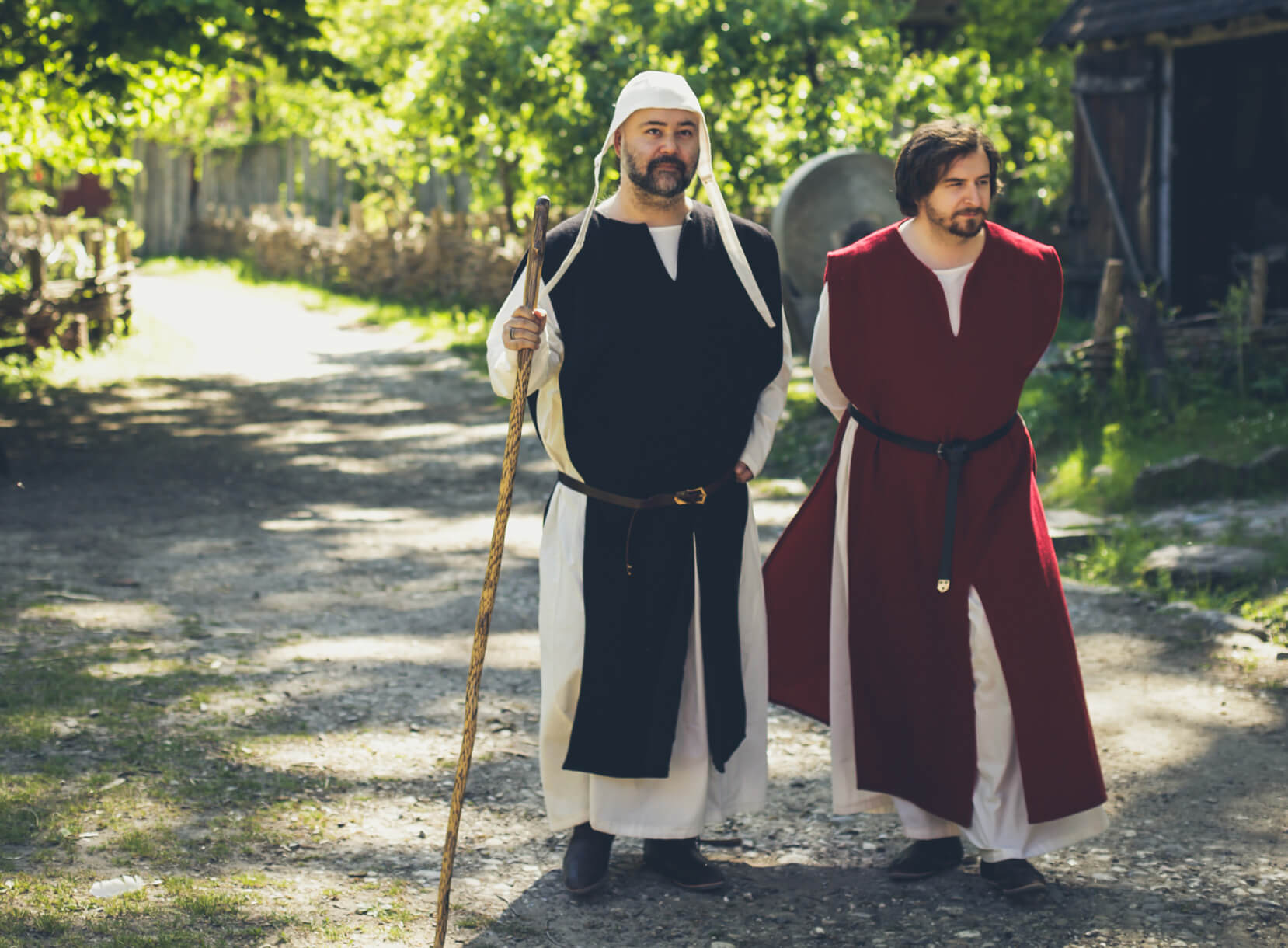 Two men in high medieval tunics, one with a staff, in historical outdoor setting.