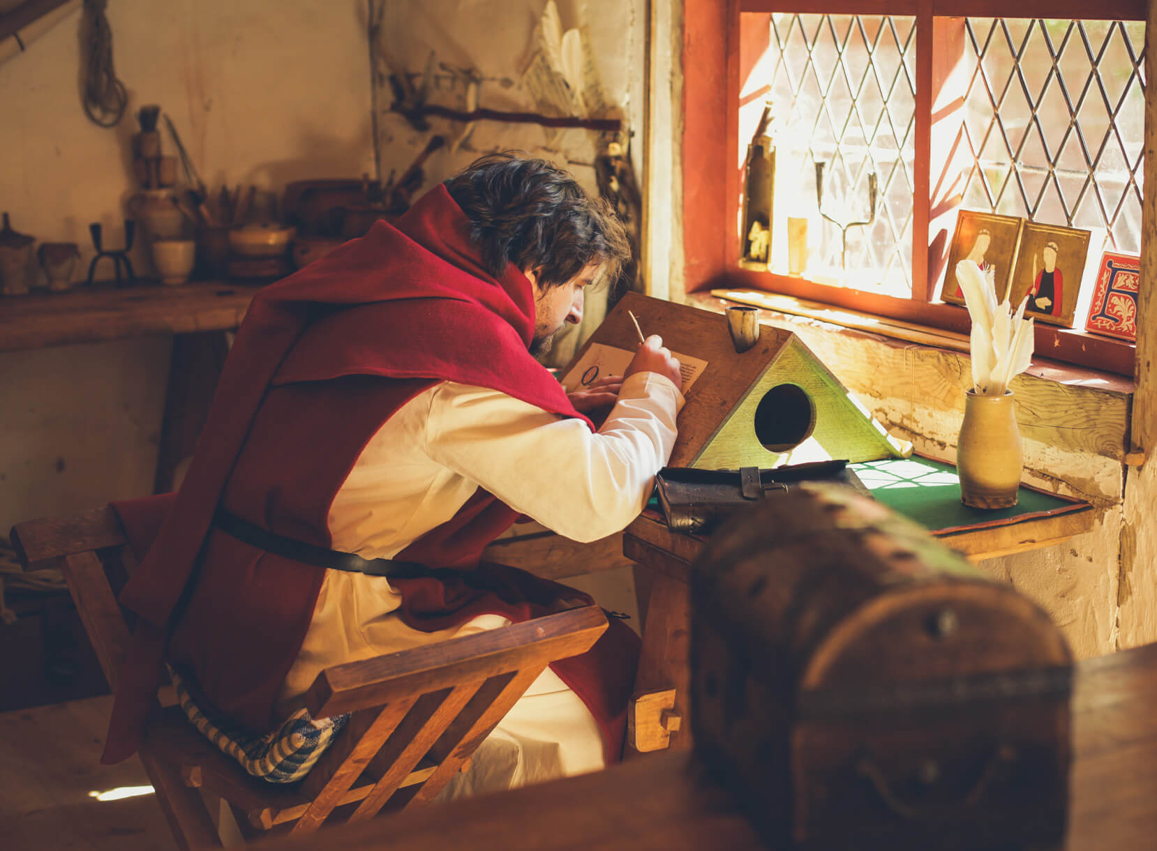 Man in red medieval hooded cape writes at wooden desk from High Medieval collection.