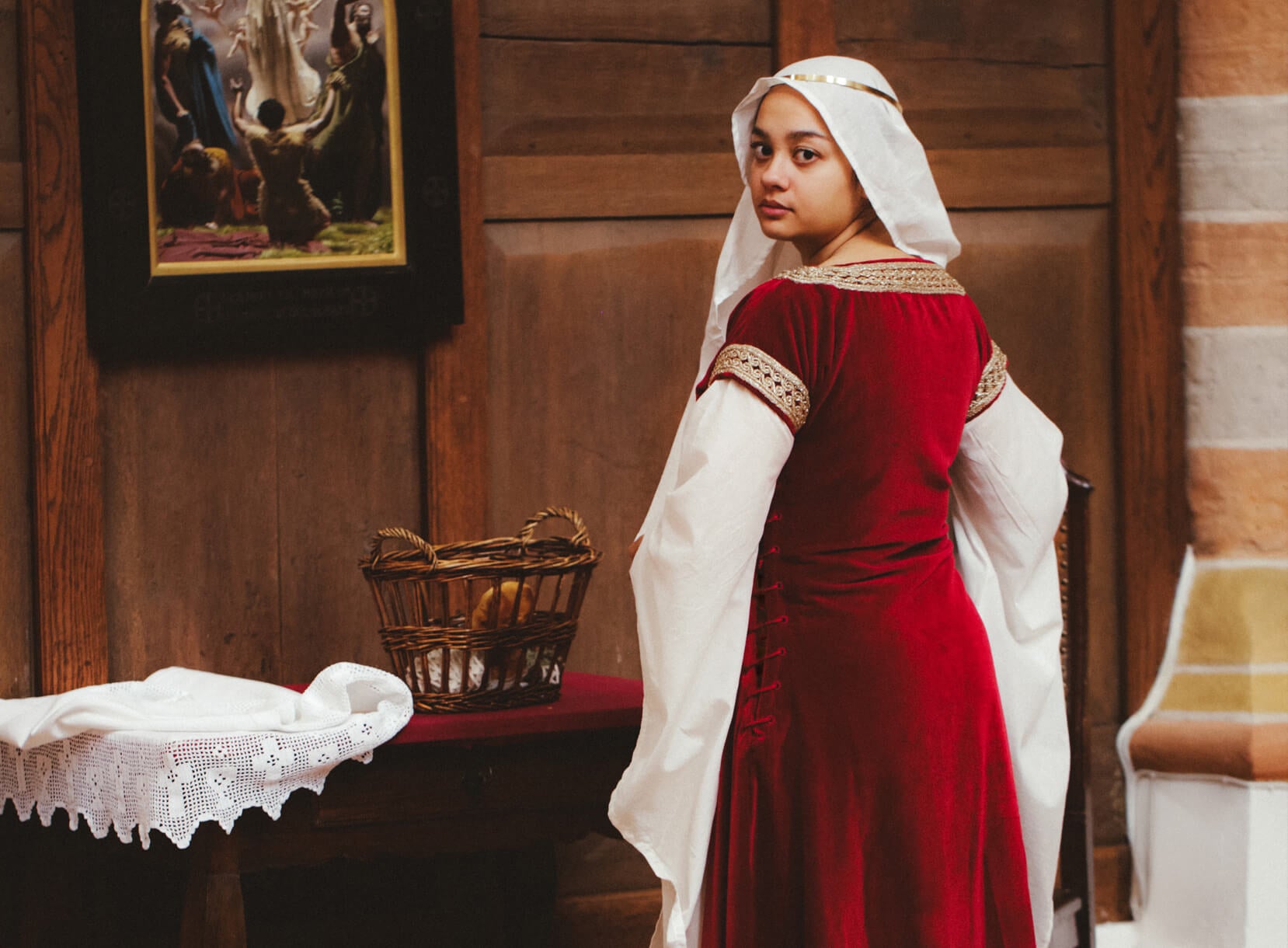 Woman in red medieval dress with white head covering, looking back from a room.
