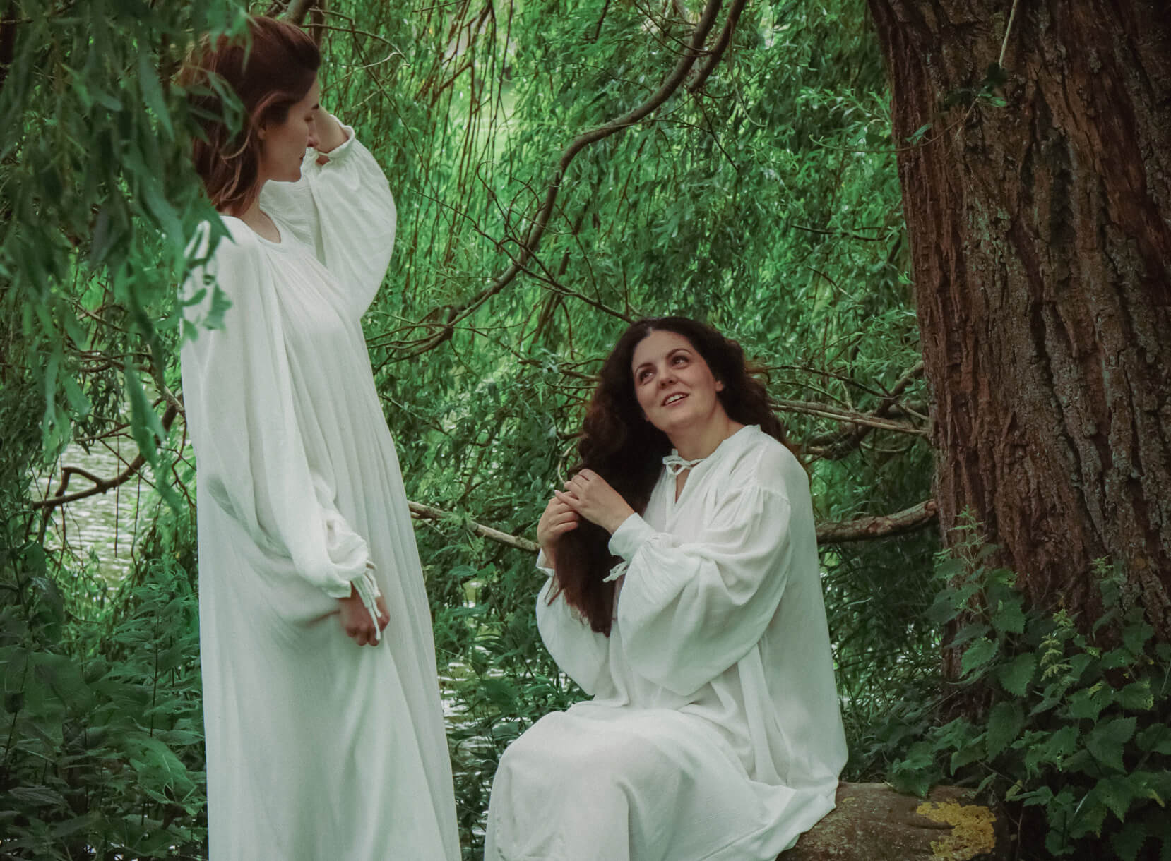 Two women in white Renaissance dresses among trees, posing for Yoremade clothing.
