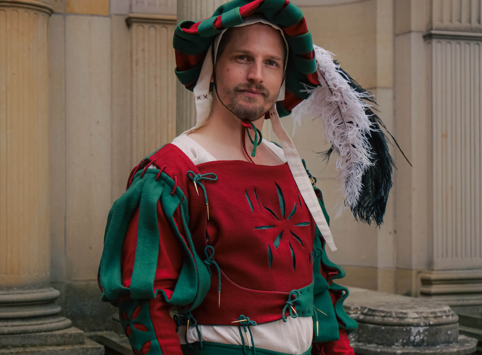 Man in luxurious red and green Renaissance Landsknecht costume with feathered hat.