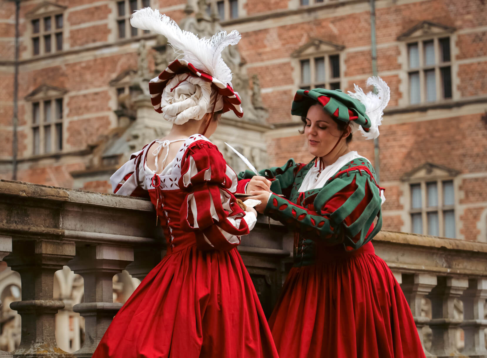 Two women in Renaissance Landsknecht costumes of red and green with feathered hats.