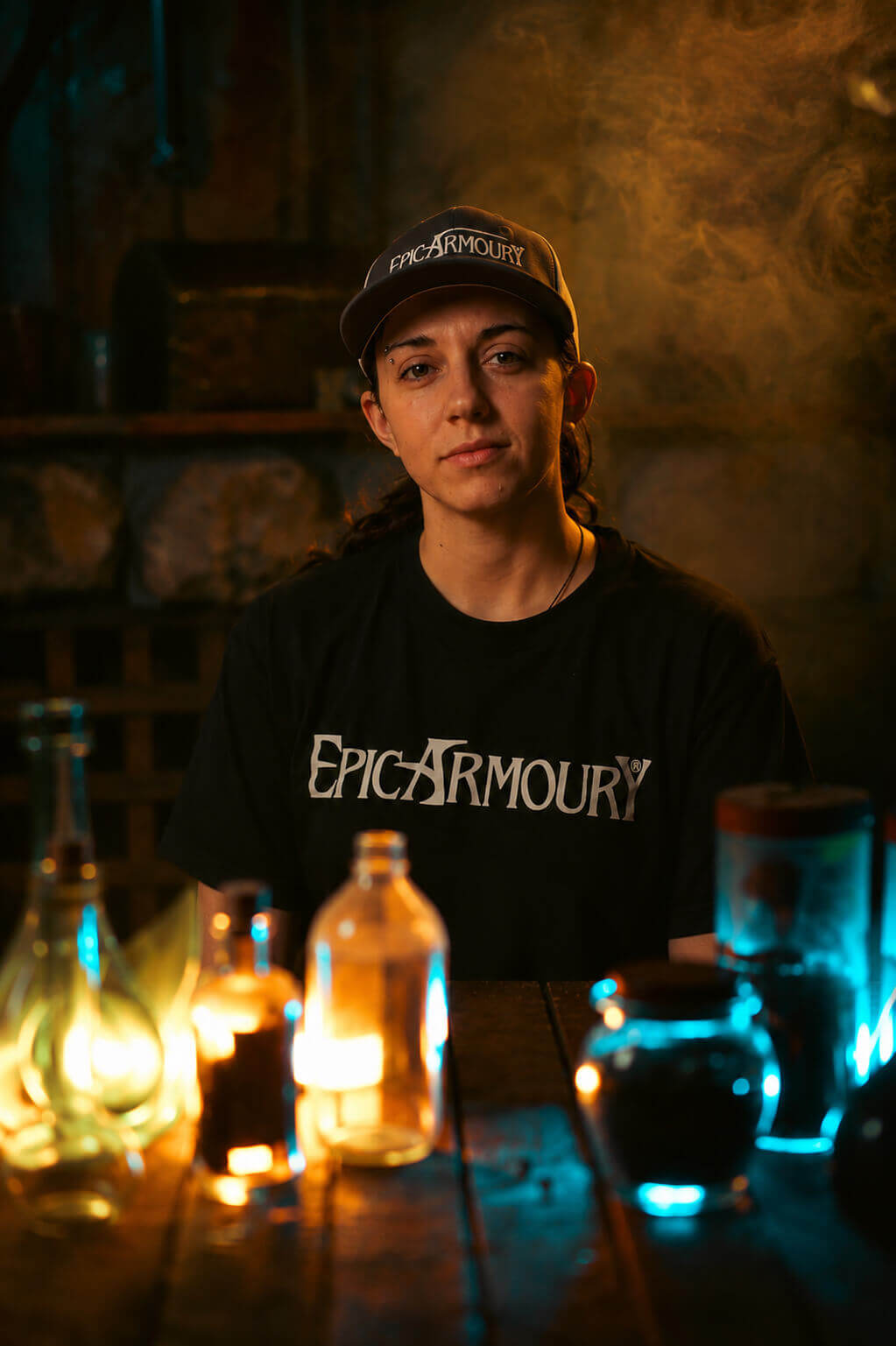 A person wearing a black t-shirt with 'EPIC ARMOURY' printed in white across the chest and a matching black cap, sitting at a wooden table with glowing bottles and jars in a dimly lit, atmospheric setting.