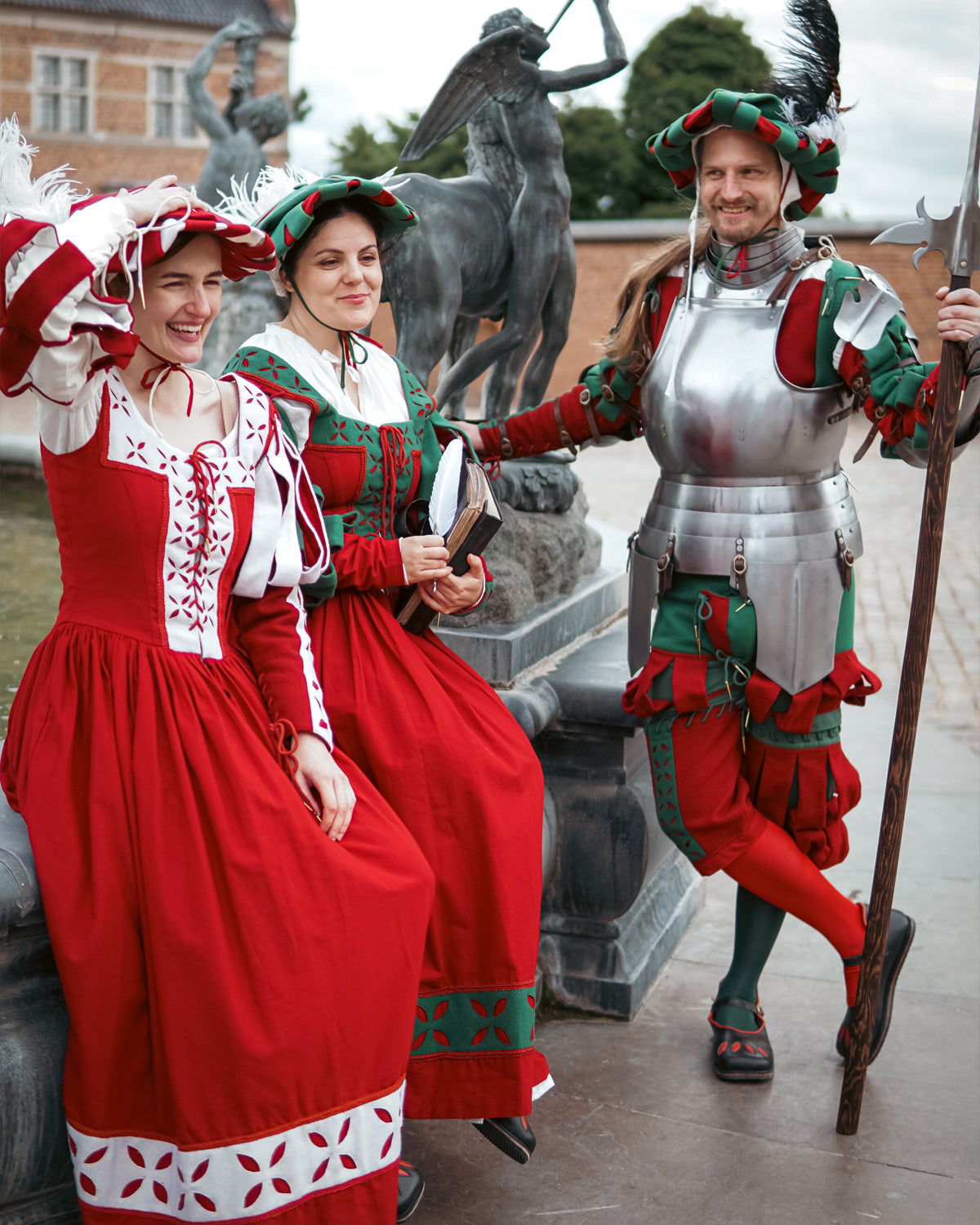 Three people in colorful medieval costumes and armor laughing outdoors.