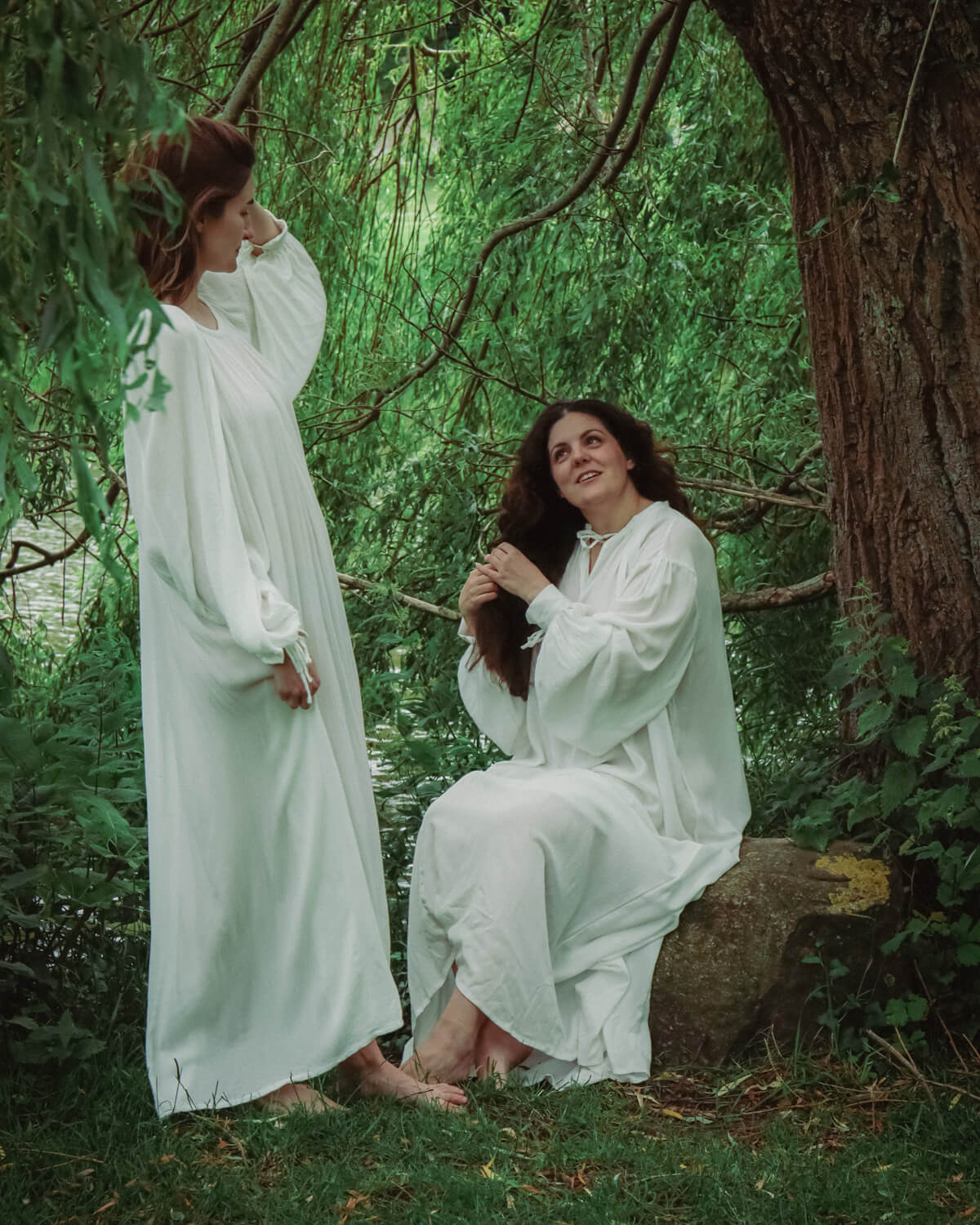 Two barefoot women in white dresses in lush green nature.