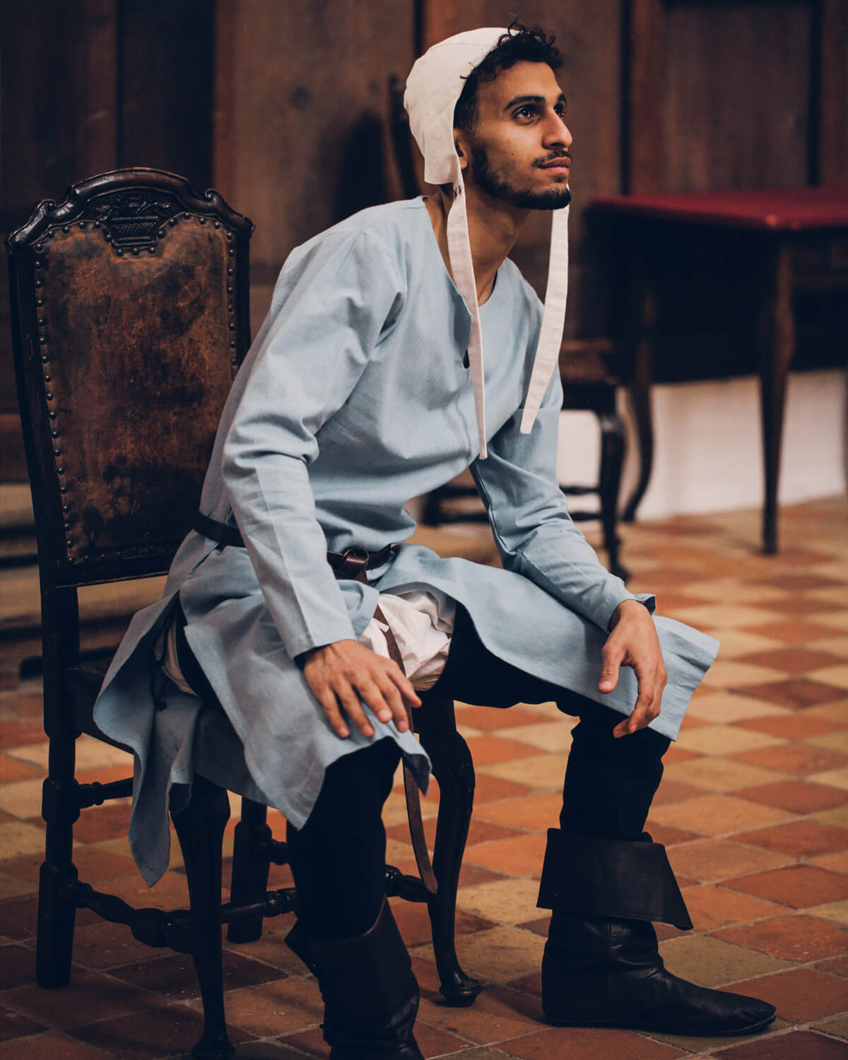 Man in light blue tunic, white coif, black pants and boots sitting on chair