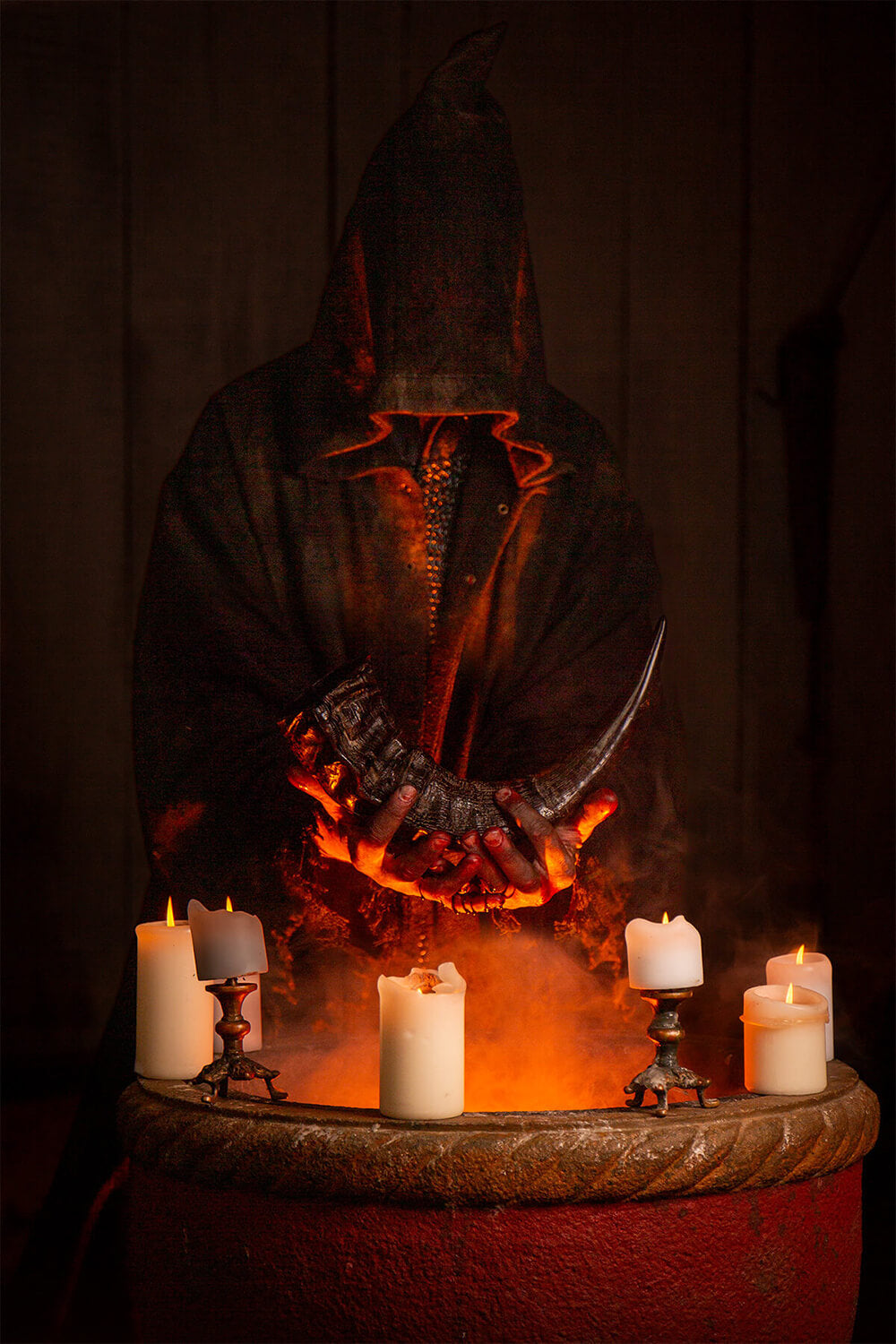 A dark hooded figure in a black robe stands behind a large red tub filled with glowing orange embers and several lit white candles, set against a dim wooden background.