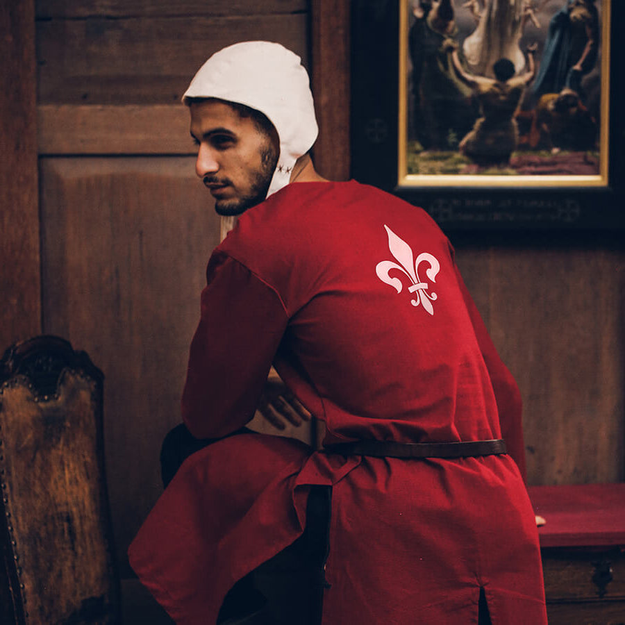 Person in a red robe with a white emblem, standing in a room with wooden walls and a painting.