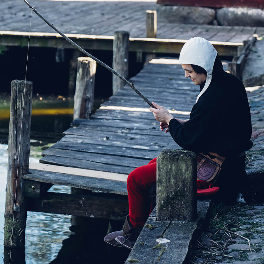 Person in a black outfit with a white hood sitting on a wooden dock, fishing.
