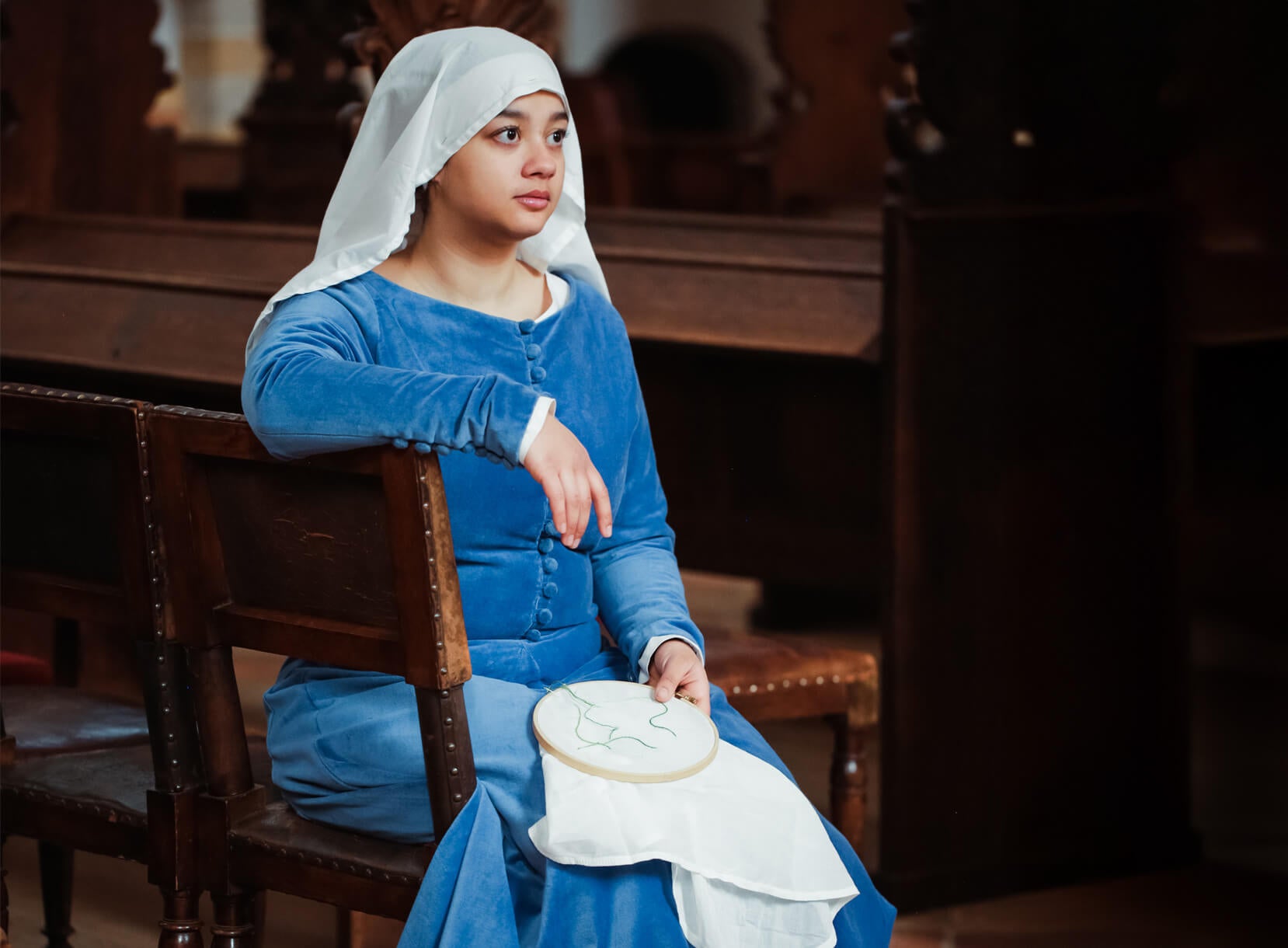 Woman in Late Medieval blue velvet dress with white wimple and embroidery hoop.