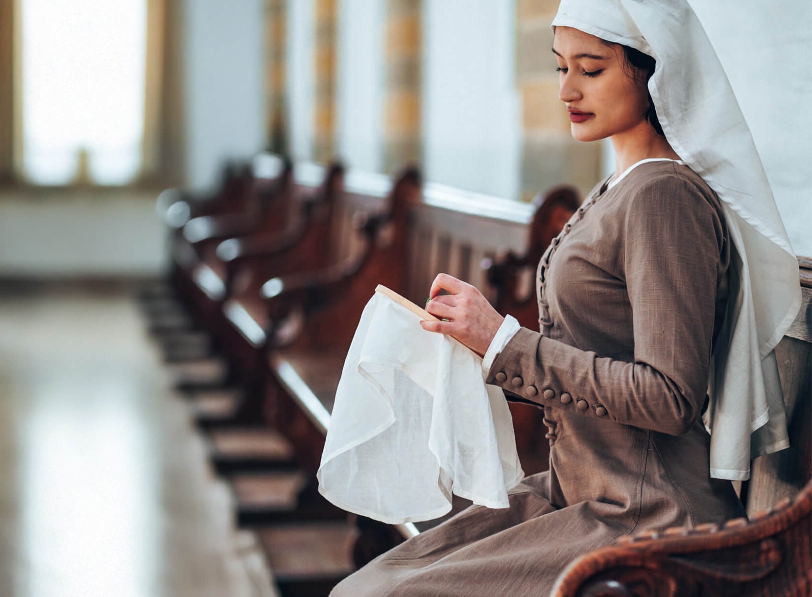 Woman in late medieval clothing sewing, with a white wimple, in a church.