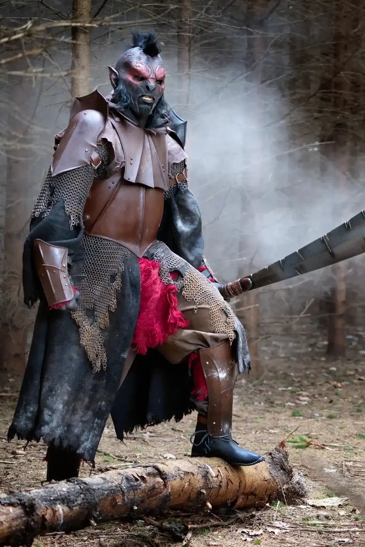 A person in elaborate fantasy armor stands on a fallen log in a wooded outdoor setting, holding a large axe. The costume includes a horned mask, dark leather and metal armor, chainmail details, and a bright red fabric accent.
