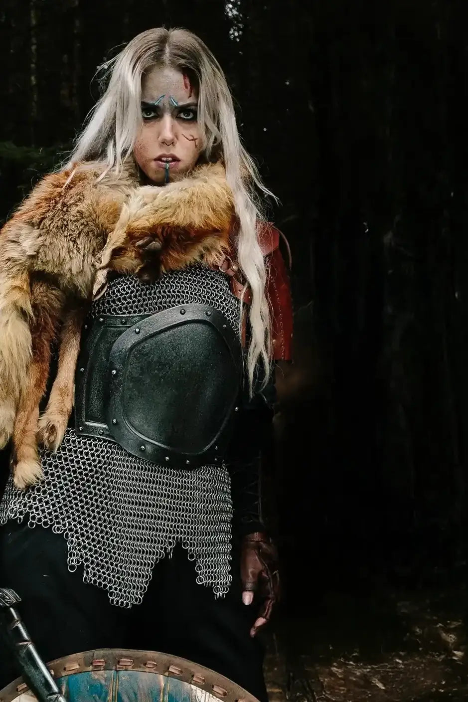 A woman in Viking-style costume stands in a dark forest, wearing a black chainmail-style tunic, a large black breastplate, a brown fur collar, and long platinum-blonde hair. She has dramatic black face makeup and a bloody lip, with a shield partially visible at the bottom.