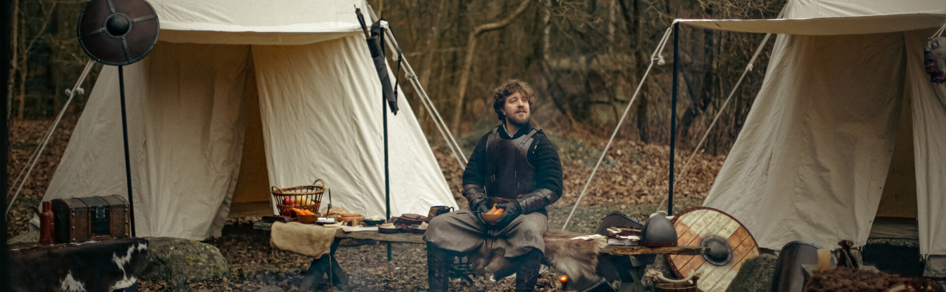 Man sitting in a forested area with two tents and camping gear around him.